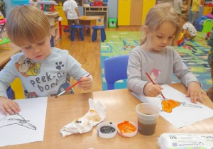 Dwoje dzieci siedzi przy stoliku w sali i maluje farbami